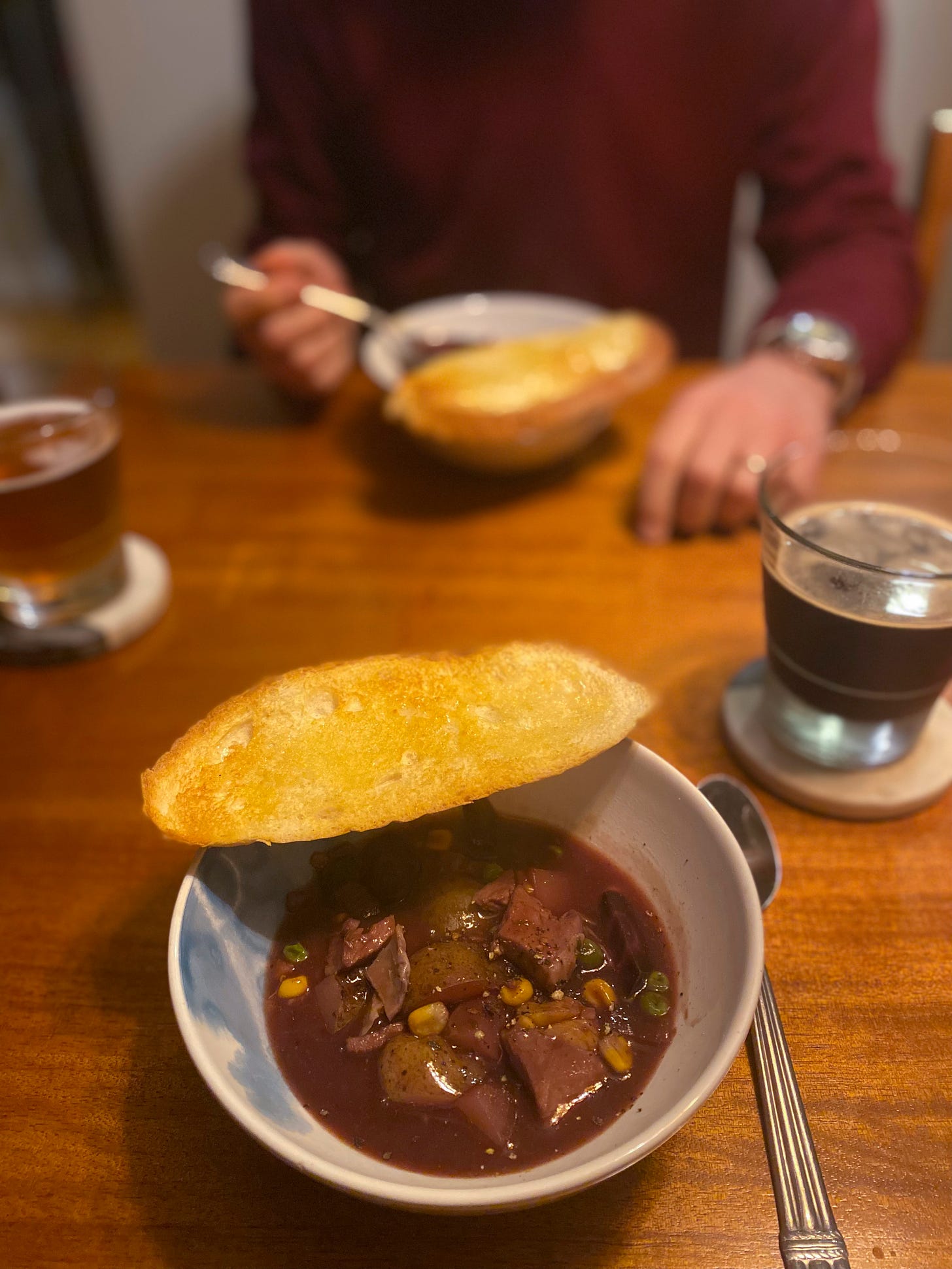 A white and blue bowl of stew with a thick, dark gravy, pieces of turkey and potato among corn and peas and onions. On the edge of the bowl is a slice of buttered toast. Across the table, Jeff holds his spoon over his bowl. Glasses of beer rest on coasters between the bowls.