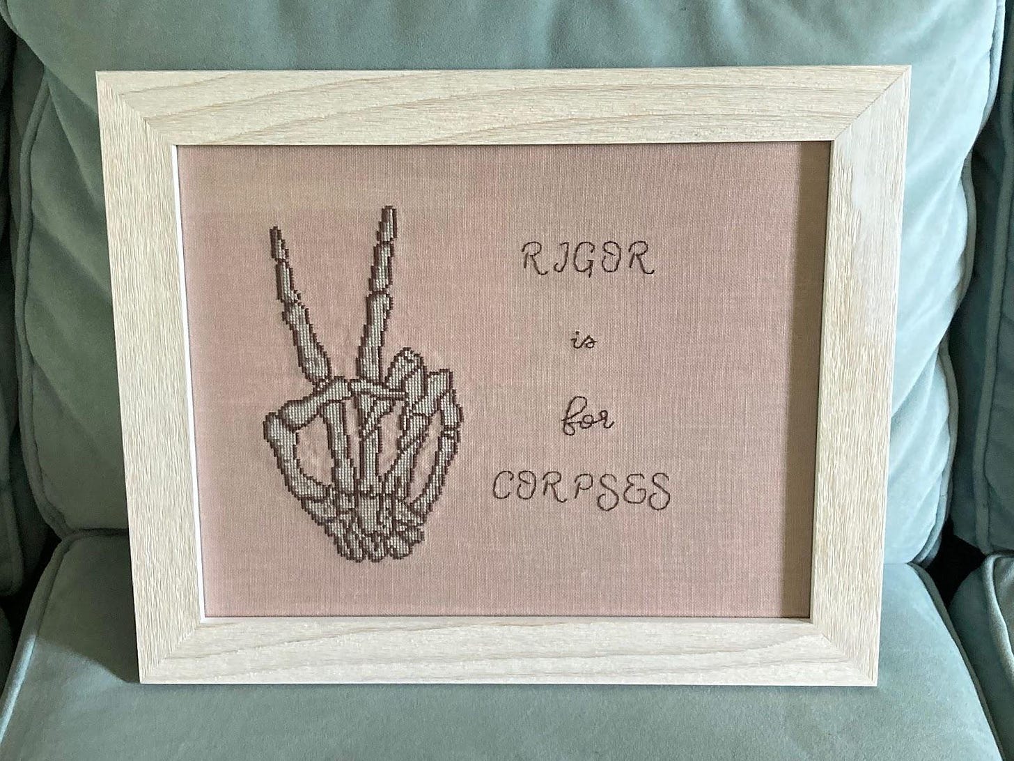 A cross-stitch with a skeletal hand and the phrase "Rigor is for corpses"