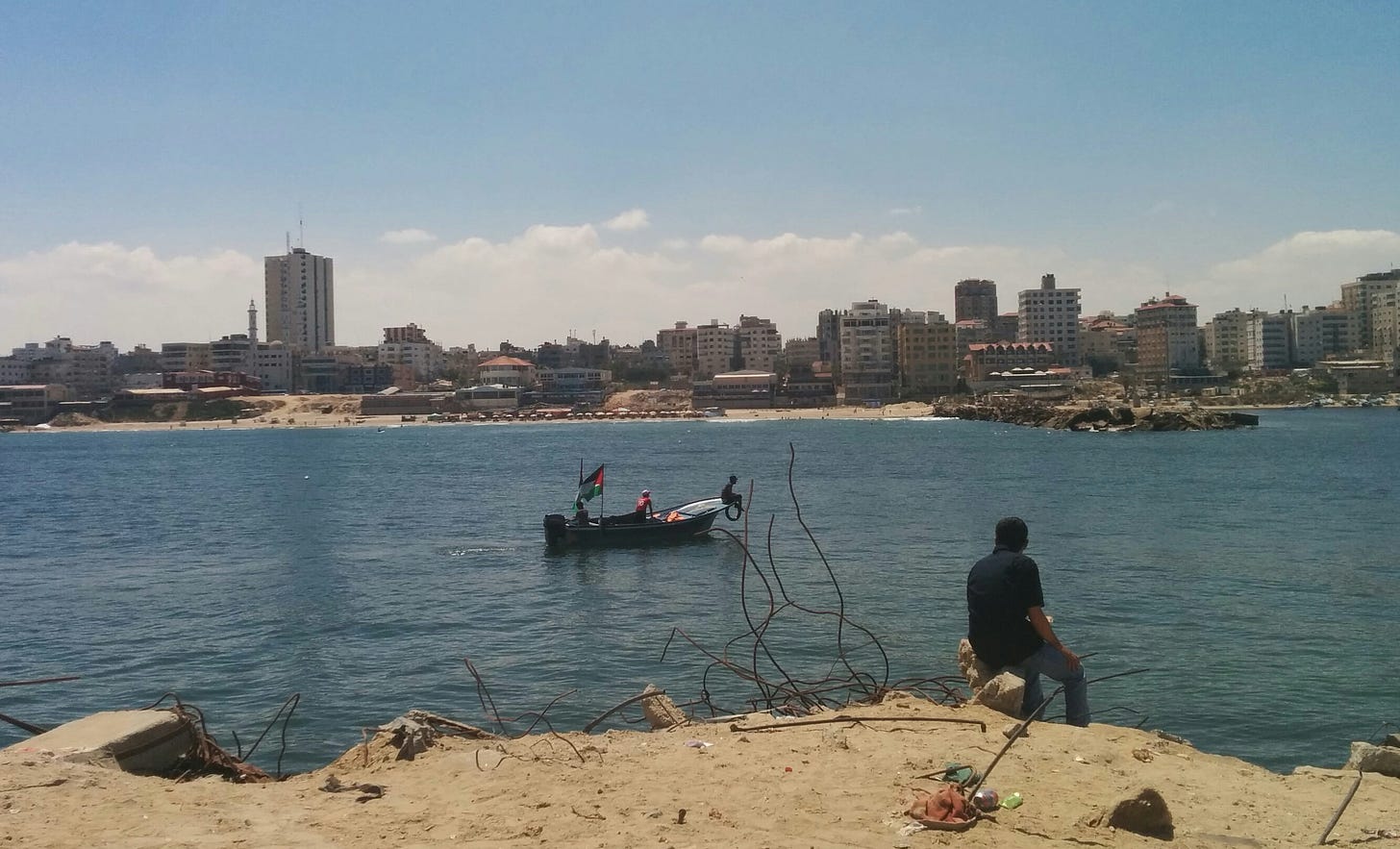 A man sits on a piece of concrete besides exposed rebar, overlooking a port with a small fishing boat flying a Palestinian flag, and Gaza City beyond