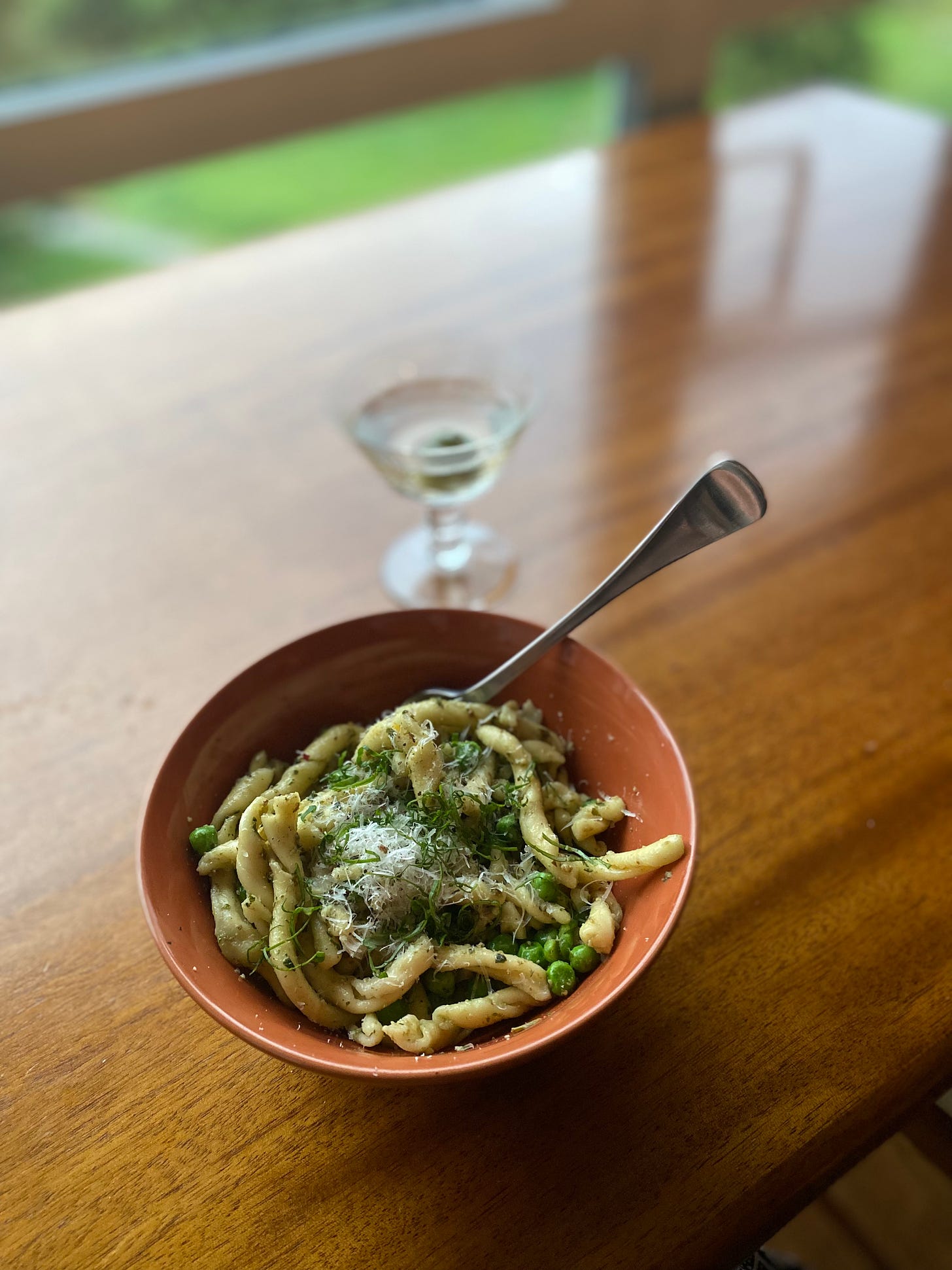 An orange bowl of medium-length twisty pasta in pesto sauce with peas, topped with basil chiffonade, grated parmesan, and cracked pepper. In the background is a martini in a coupé glass.