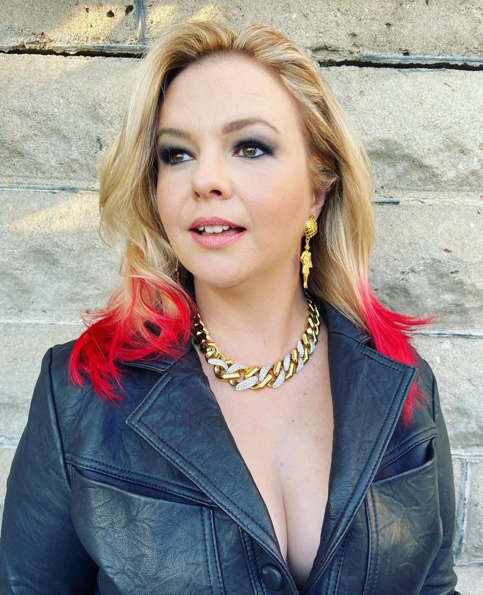 Amber poses wearing a lowcut black jumpsuit. Her blonde hair has bright red tips.