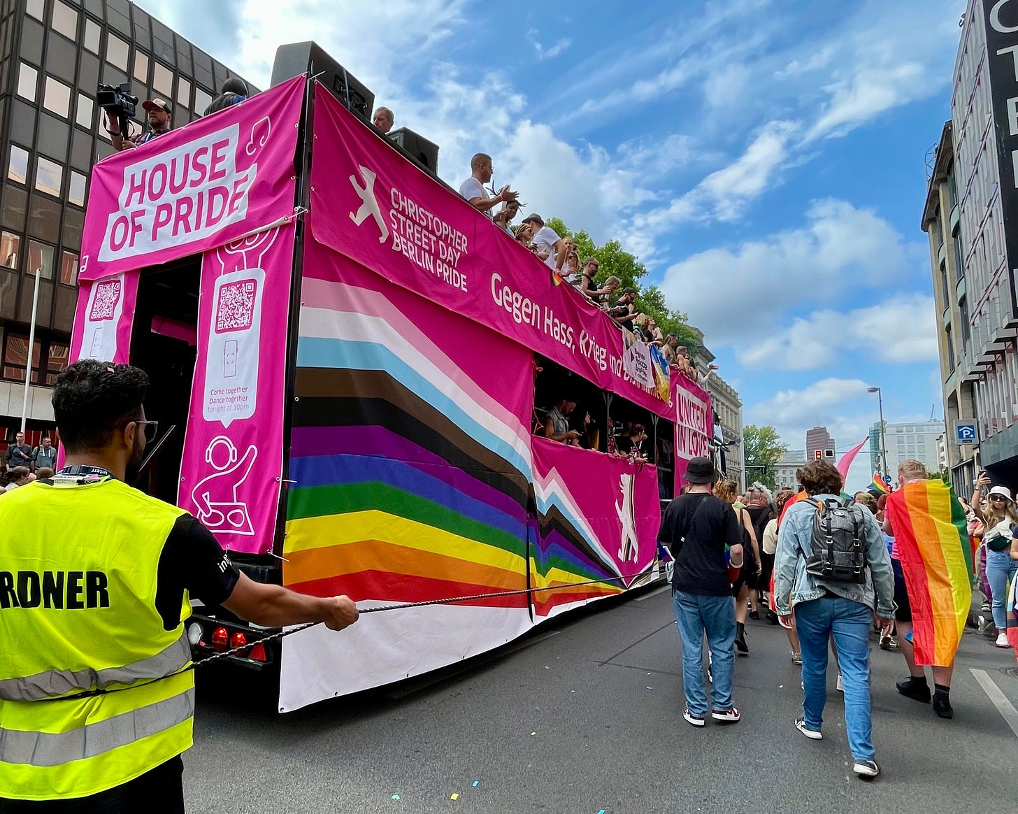 A vibrant float at Berlin CSD 2023 adorned with a large Progress Pride flag and pink banners reading "House of Pride" and "Gegen Hass, Krieg und Gewalt" (Against hate, war, and violence). A crowd follows the float as it moves down the street under a sunny sky.