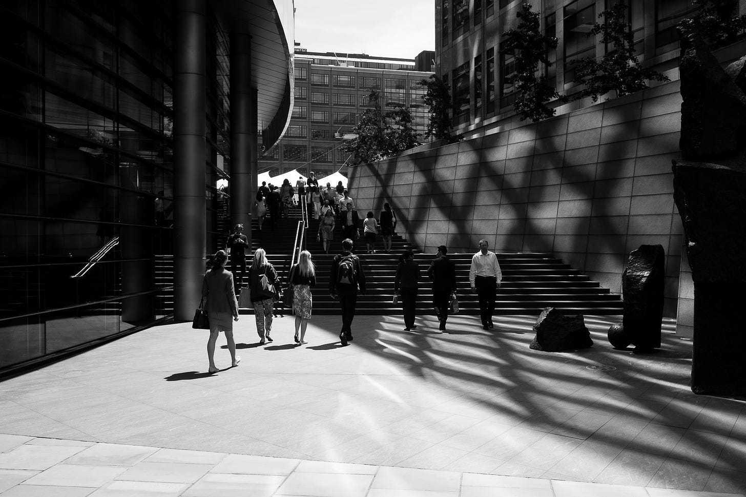 London, 2015. People walk past a building. On the wall across from the building there is a criss-cross shadow.