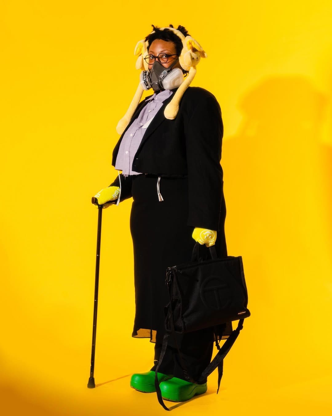 A black woman stands against a yellow background wearing a respirator mask, gloves, and formal suit. She holds a cane in her left hand and a briefcase in her right hand, while looking towards the camera. 