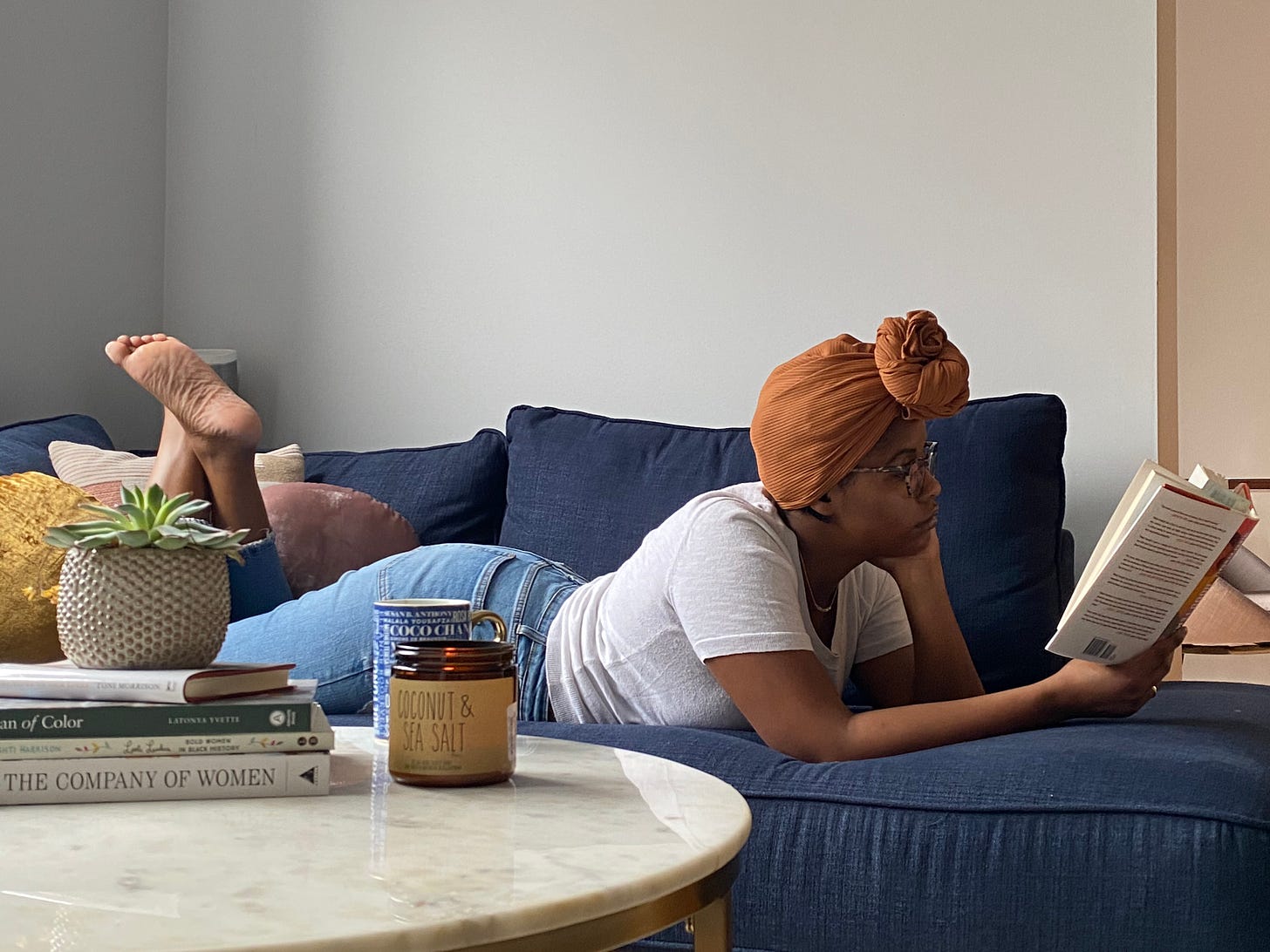 Black woman lying down reading a book on a blue sofa