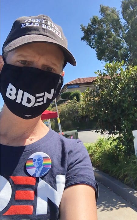 Photo of the author wearing a black face mask with BIDEN across the front. It covers most of the lower part of her face. She is also wearing a black ball cap and a navy t-shirt with BIDEN on it and a photo button of Joe Biden