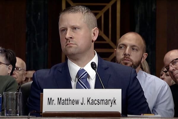 A still image taken from a video feed of a Senate judicial hearing shows Matthew Kacsmaryk in a dark blue suit sitting at a table with a microphone and a name placard. Several audience members are in attendance behind him.
