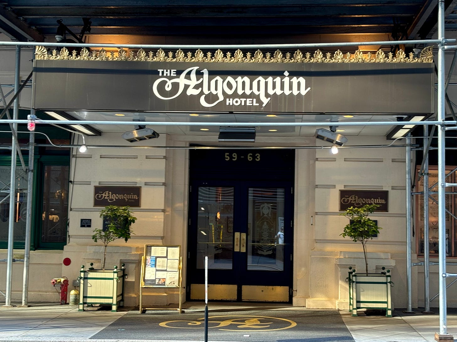 The awning and entrance of the Algonquin. There are names plaques on either side of the door and a menu board for the restaurant. The windows on either side are full of historical odds and ends.