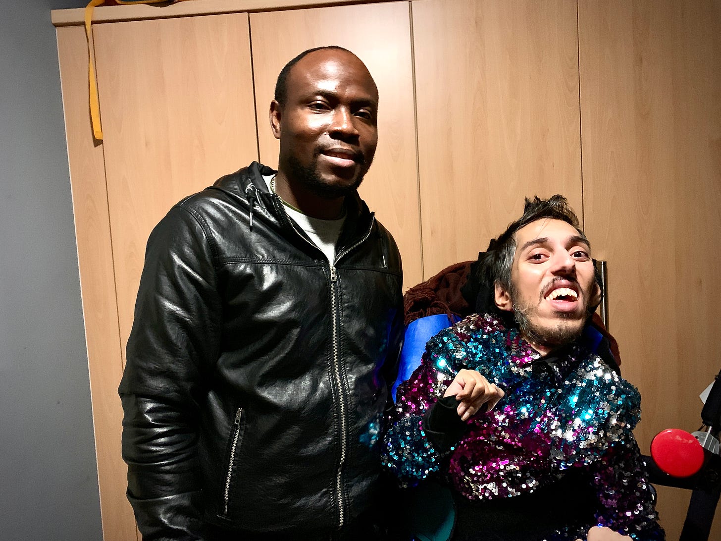 IMAGE DESCRIPTION 1: Photo from 9 December 2019 of Sulaiman (a wholeheartedly Disabled AF and British-Pakistani man) sitting in his power wheelchair in his room next to his carer Albert (a tall, badass Black man from Ghana, West Africa) after a night on the town. Sulaiman is wearing his bright rainbow-coloured jacket and Albert is wearing a faux black leather jacket. They’re both smiling and are joyful. DESCRIPTION ENDS.