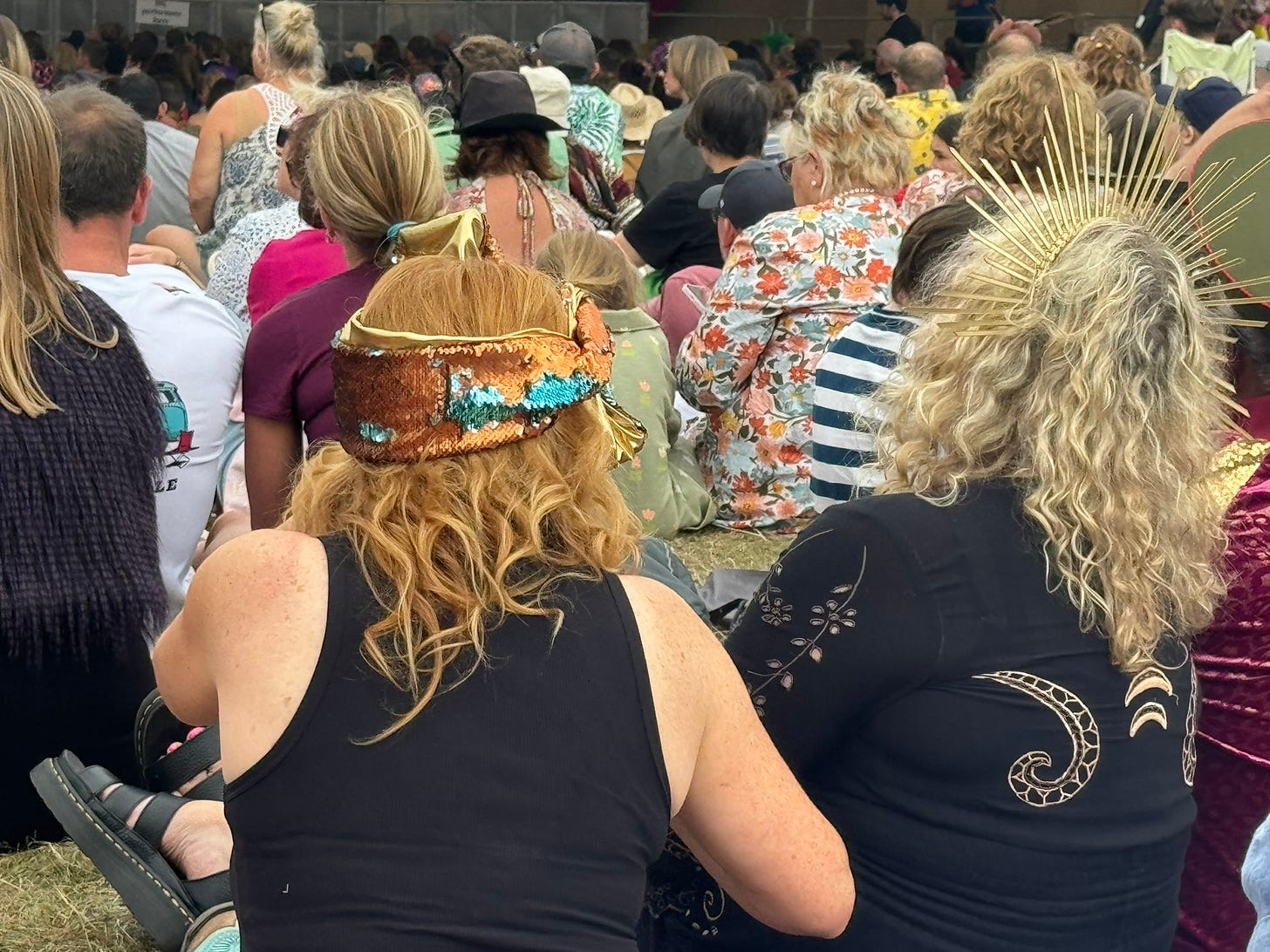 the backs of audience sat on ground.  2 ladies closest have interesting headware.  A bronze bandana and the other lady is wearing a gold spiky band.