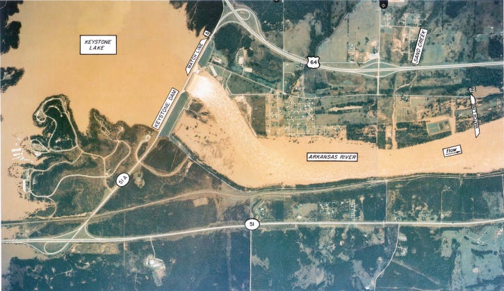 The vintage aerial photograph shows the mud-orange flood waters of Keystone reservoir, feathering across dark green surroundings, frothing as it passes through Keystone Dam and down the swollen Arkansas River.