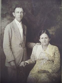 Portrait of a gaunt man in a light colored suit standing beside his sitting wife wearing a floral pattern dress.