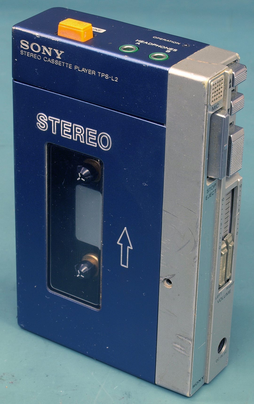 Close-up of the original Sony Walkman TPS-L2, a blue and silver portable cassette player from 1979. The device features dual headphone jacks, a large orange HOT LINE button, and metallic volume sliders along the side. Close-up of the original Sony Walkman TPS-L2, a blue and silver portable cassette player from 1979. The device features dual headphone jacks, a large orange HOT LINE button, and metallic volume sliders along the side.