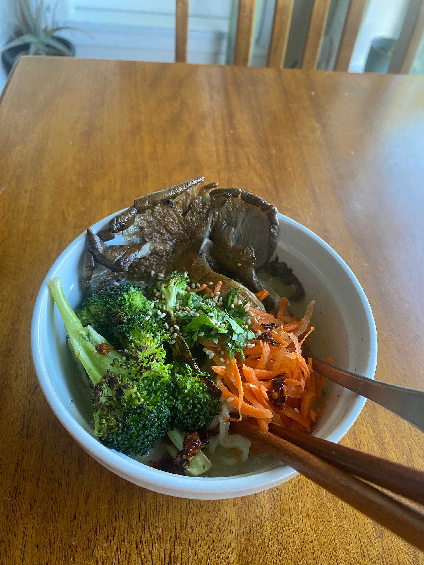 The soup described above, a greenish-yellow broth filled with noodles and topped with seared broccoli and oyster mushrooms, shredded carrot, chopped cilantro, and sesame seeds. Chopsticks and a spoon stick out of the lower right side of the bowl.