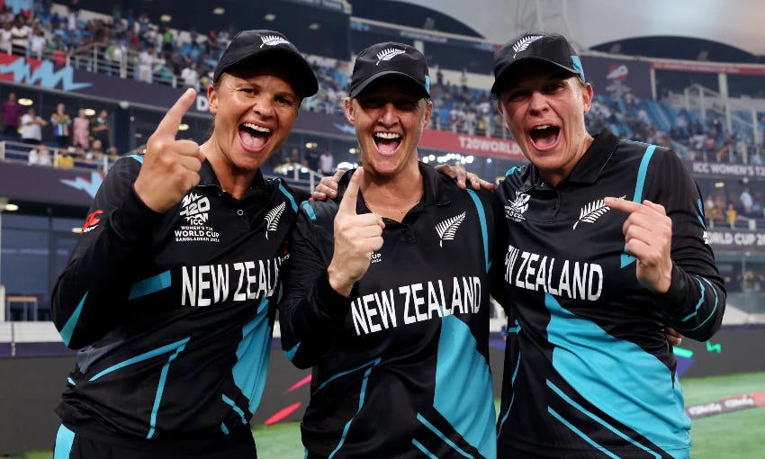 Three New Zealand women's cricket players, Suzie Bates, Sophie Devine and Lea Tahuhu, celebrate on the field while smiling and pointing. They are wearing black and teal uniforms with "New Zealand" printed on them. A stadium full of spectators is visible in the background.