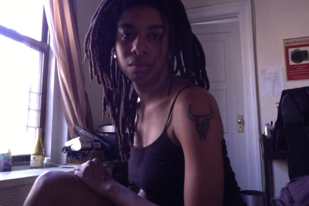 Self Portrait of young black woman with dreadlocks staring straight into the camera. Her left arm shows a prominent bull tattoo.