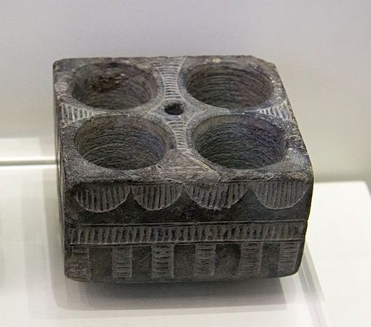 Square dark grey stone libation table with four circular hollows for receiving liquid offerings. There are shallow geometric designs incised on the sides and the top.