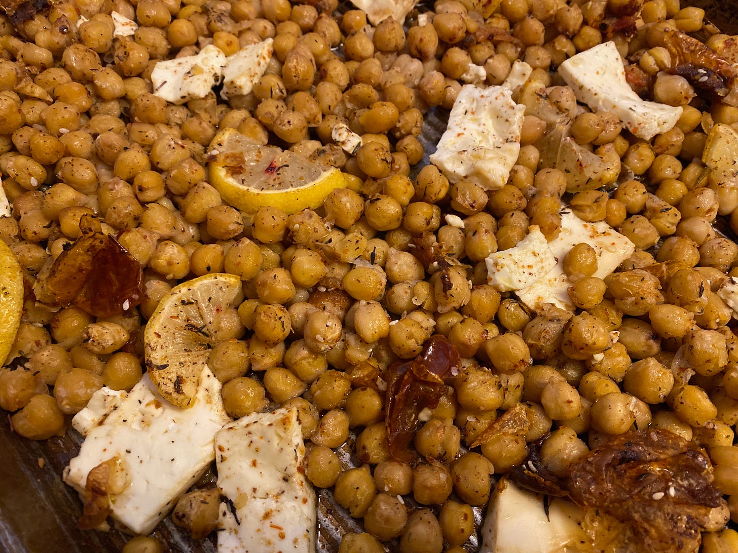 A tray of roasted chickpeas, with slabs of feta, dried tomatoes, and sliced lemons nestled among them.