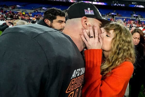 Travis Kelce, left, wearing football pads with an AFC Champion T-shirt and hat that says Super Bowl, kisses Taylor Swift on the field after a game.