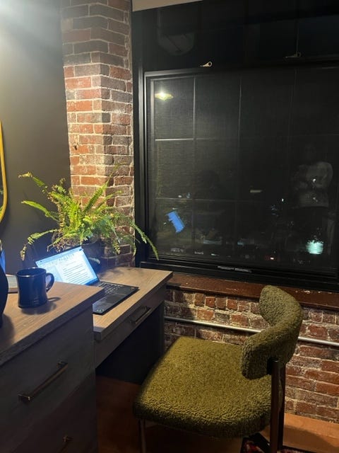 photo of my morning writing spot in my hotel room: brick wall, black window, underwatered fern, and my laptop and coffee