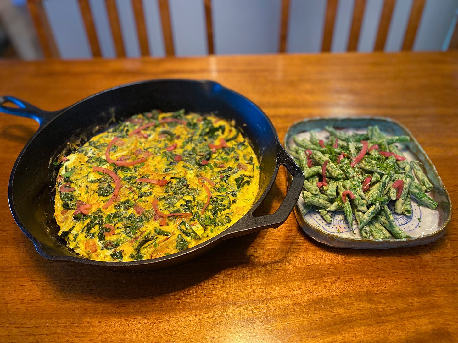 A large cast iron pan with the frittata described above, with parsley, dill, and pickled red onion over top. Next to it is a small blue ceramic dish with the green bean salad described above, also garnished with the herbs and onion.