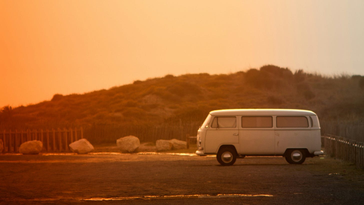 A hazy orange sunset with a rustic VW van parked on an open road.