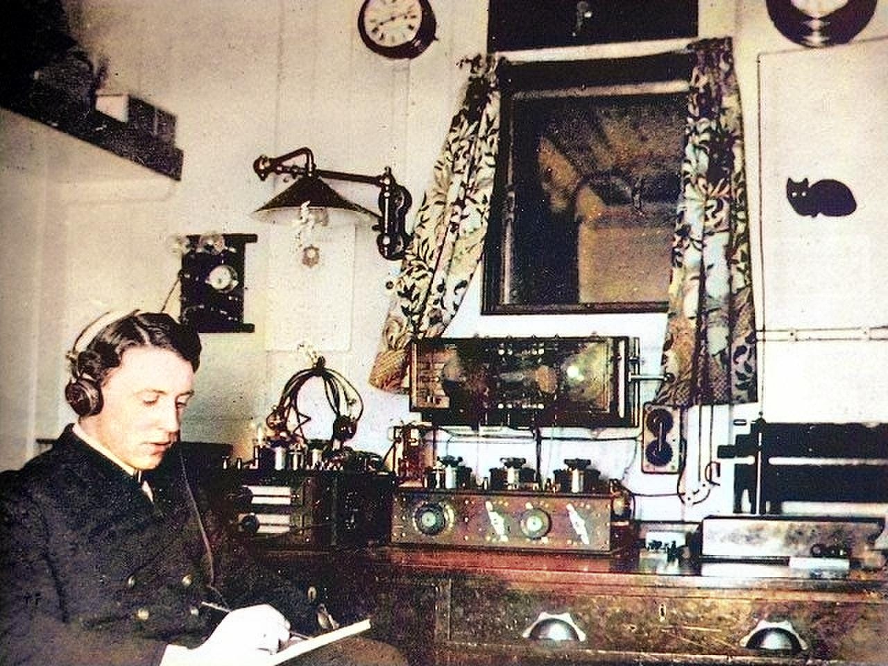 A young man wearing headphones in a lace-curtained room with radio equipment: everything old-fashioned, naturally, as the photo was taken in 1913.