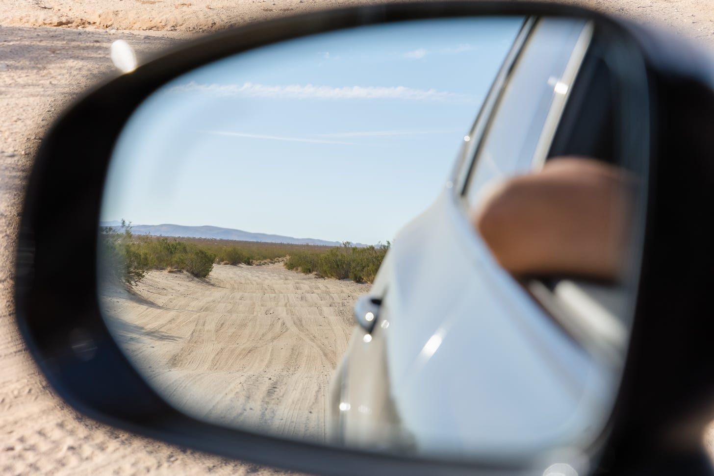 Daytime image of a rearview side car mirror, viewing a wavy, sand road in the middle ground, an elbow perched on a car window in the foreground, and mountains in the background.