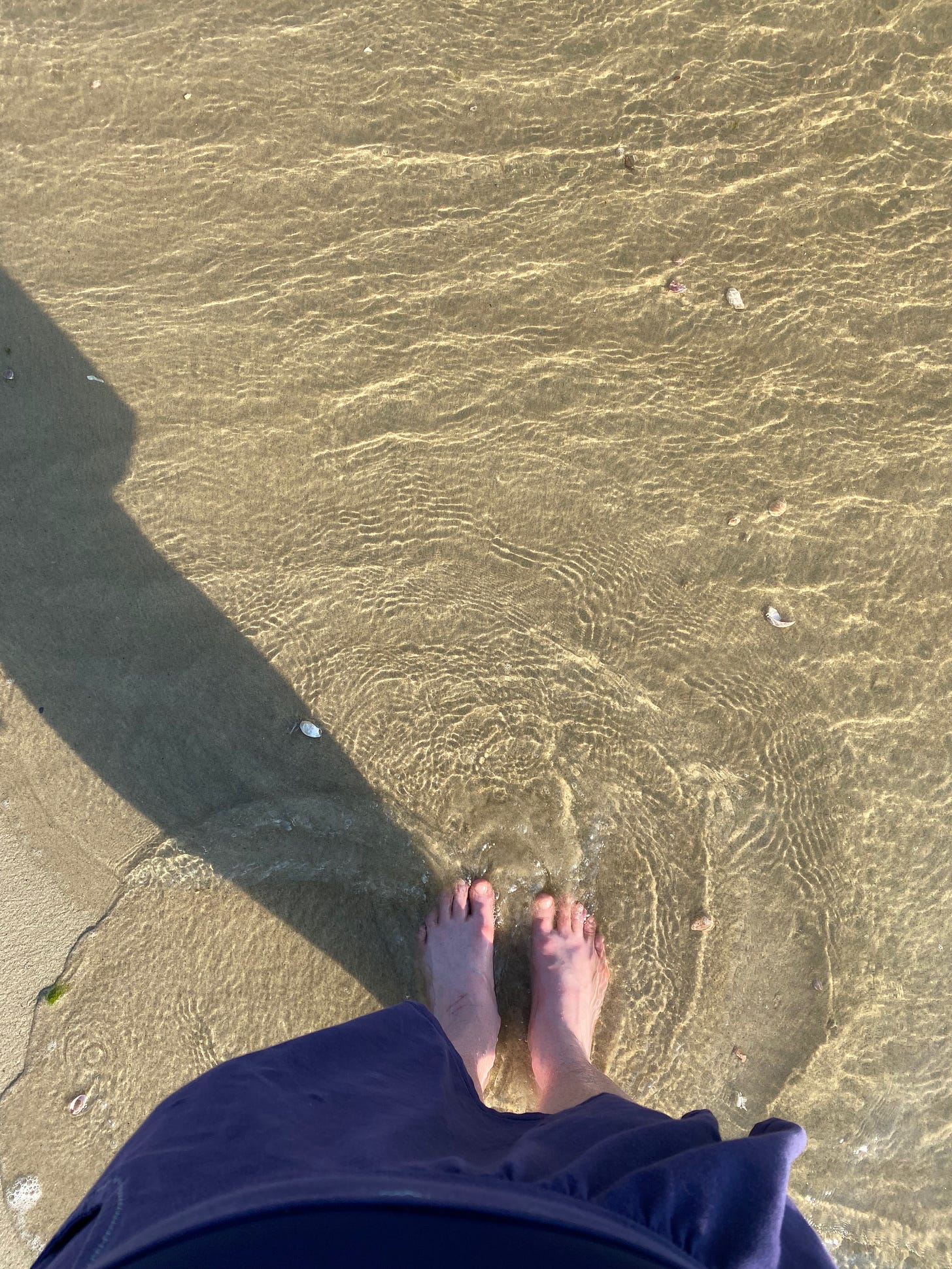 A view from above of my bare feet in a swirling wave of clear water on the beach. I’m wearing a short purple skirt and my legs make a long dark shadow.