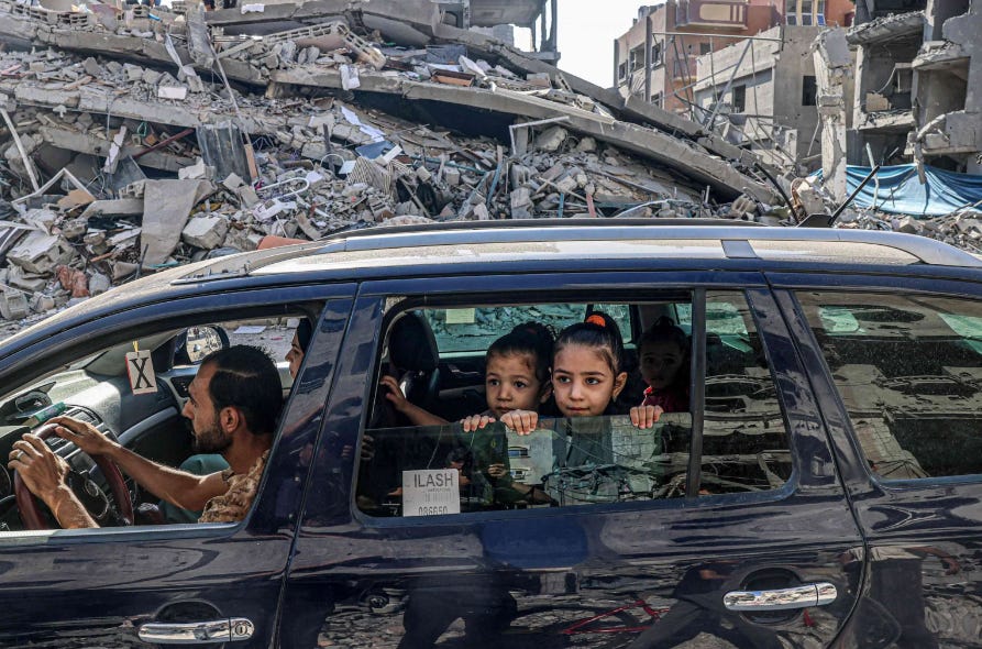 Three infant aged Palestinian girls look out the right rear passenger window as their mother sits in the front passenger sit as her father drives through the rubble of bombed out buildings in Rafah in Southern Gaza on October 12, 2023. Photo by Said Khatib via AFP.