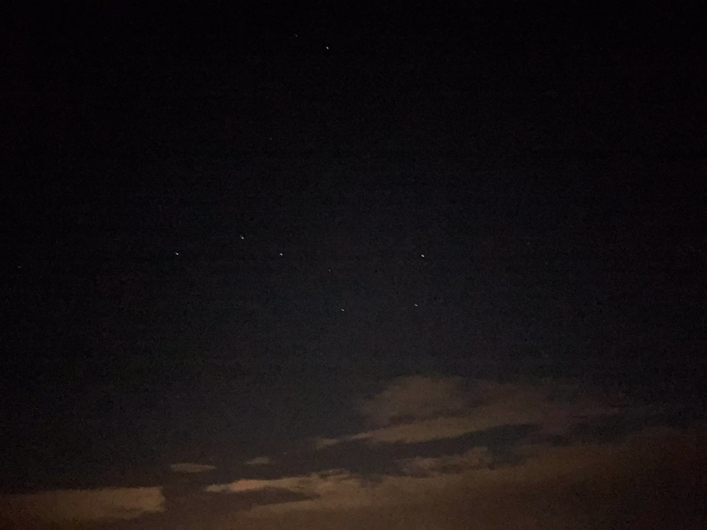 The night sky appears as a dark black space fading lighter at the bottom, with several clouds along the bottom of the photo. Above the clouds, the Big Dipper constellation can be seen as a series of six tiny bright dots. 