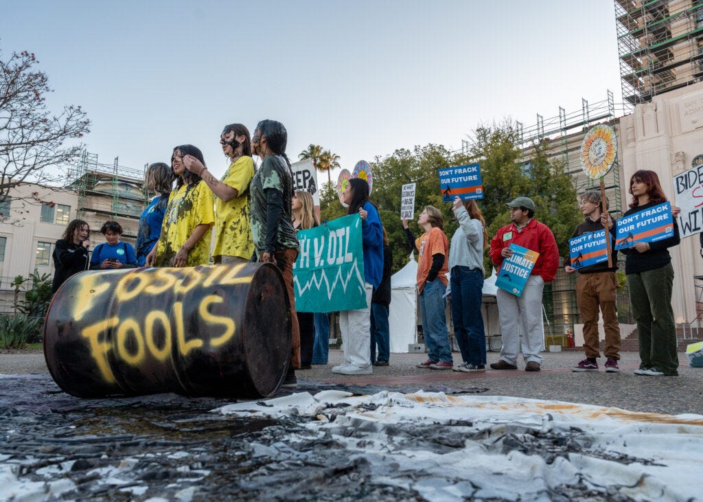 Youth members holding signs and youth members covered in oil in front of an oil barrel that reads "fossil fuels"