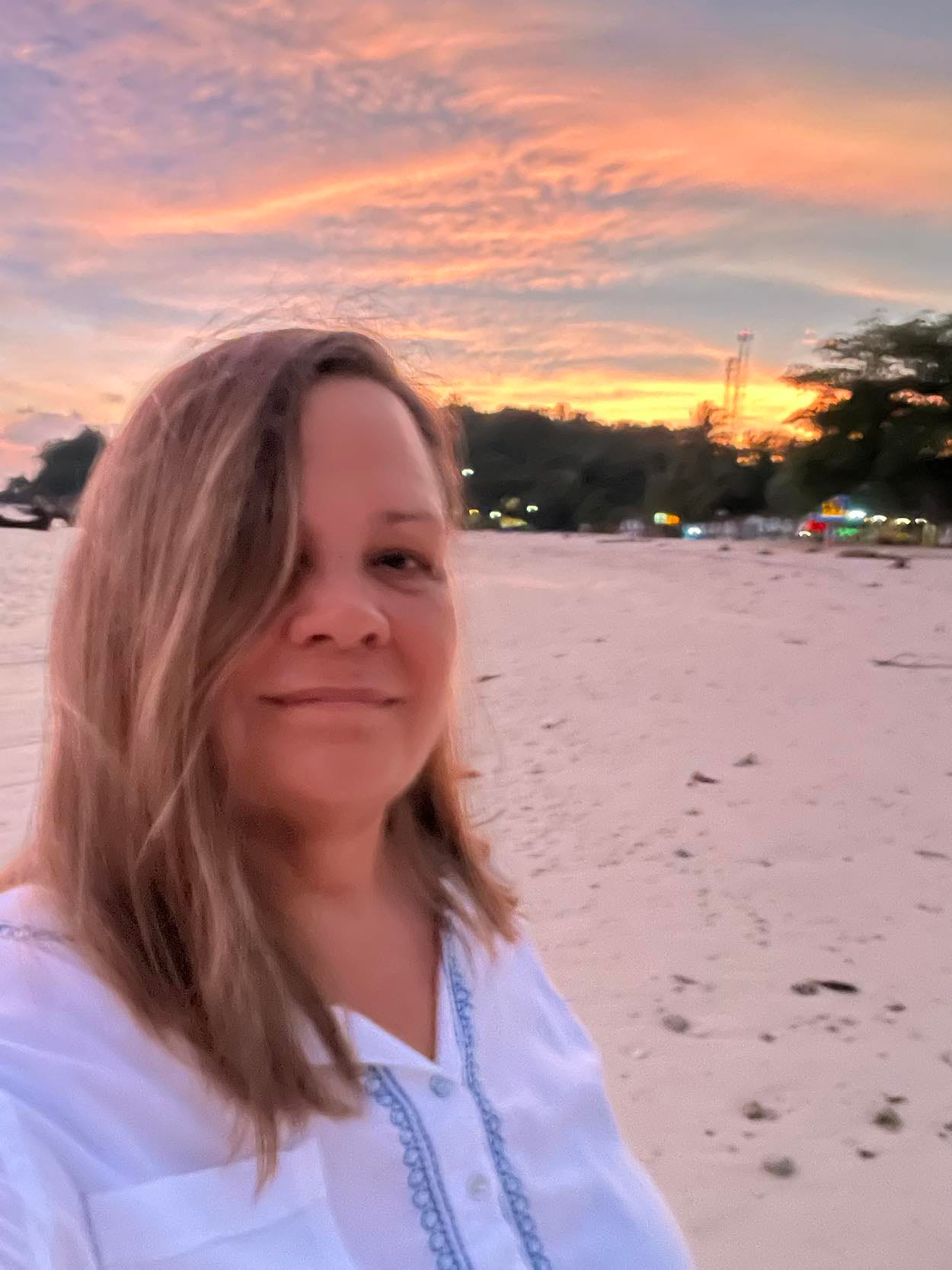 a woman on a sandy beach in front of a soft and colorful sunset. She is smiling contentedly.