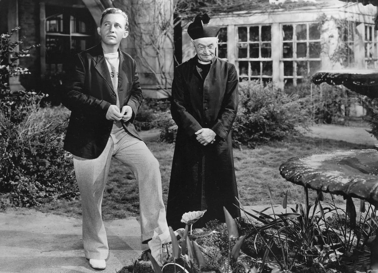 Bing Crosby (left) in casual wear and Barry Fitzgerald in an old-fashioned Catholic Prisest’s surplice and cap in Going My Way (1944). They’re in the garden of the priest’s home. Corsby looks up and out of shot with a serious expression. Fitzgerald look down at a flower bed contemplatively