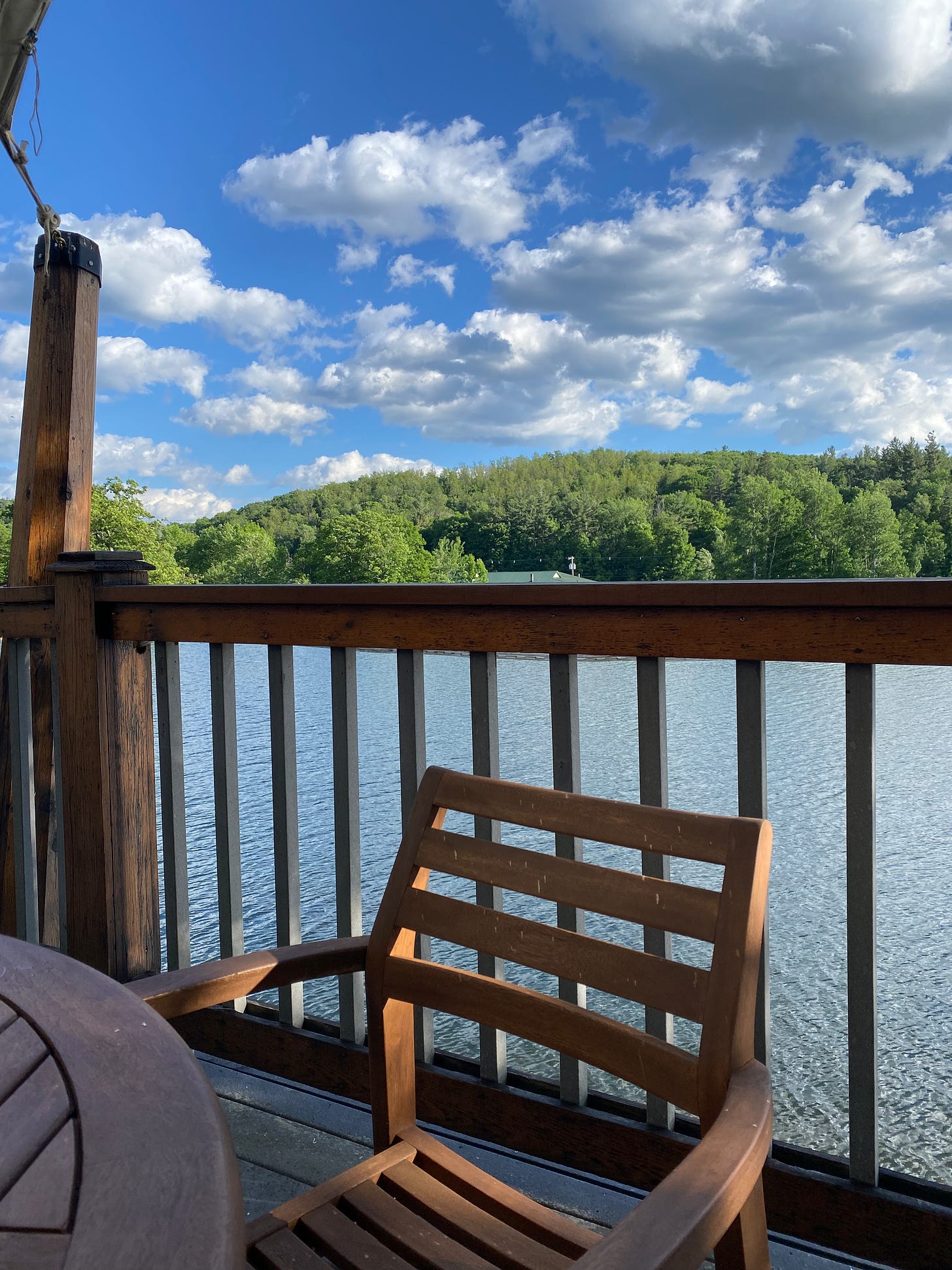 A wooden chair next to a railing on a porch overlooking Ashfield Lake. The sky is bright blue and dotted with clouds; the hills in the background are bright green.