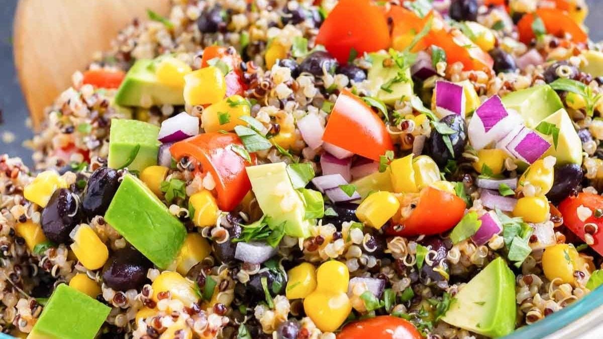 A colorful quinoa salad mixed with black beans, corn, avocado, diced red onions, cherry tomatoes, and herbs in a clear bowl with a wooden spoon.