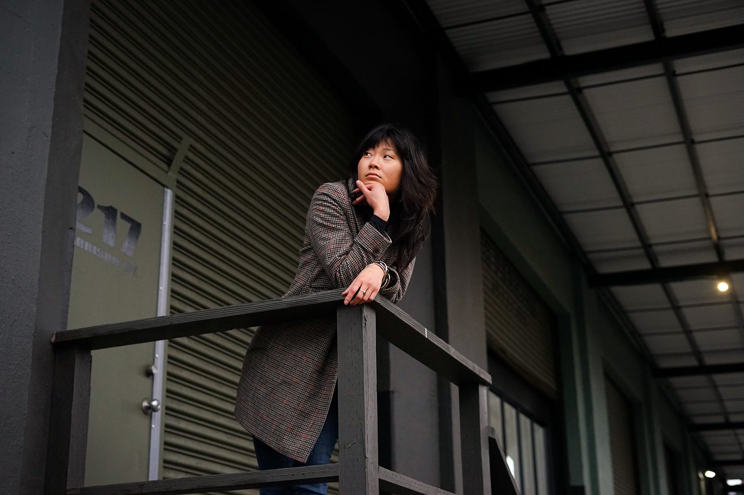 An asian female leaning over a guardrail outside a warehouse, chin resting on her hand gazing out into the distance