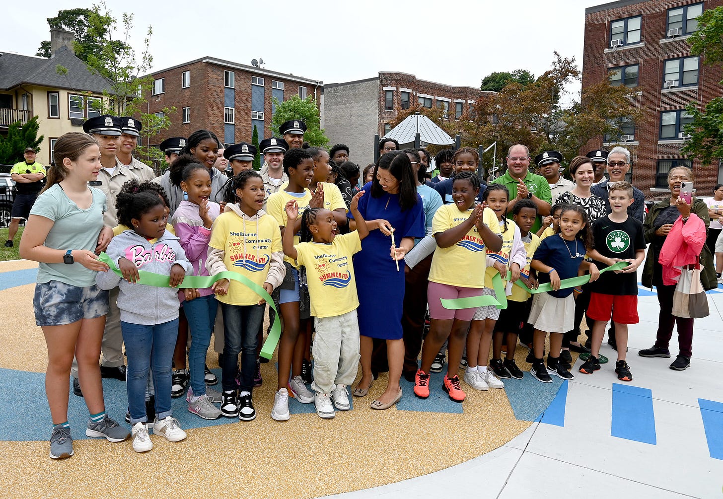 Mayor Wu and Parks Commissioner cut a green ribbon to officially open the new Walnut Park Play Area alongside about a dozen young people, members of the Boston Police Cadet program, and community members