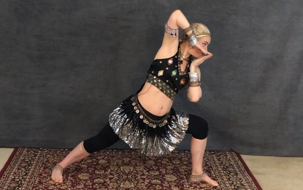 The author, blonde today, in a black and silver costume with mirrors, coins, ankle bells, and metal bangles. Lunged in a deep bow-and-arrow stance with angular arms and a piercing gaze.