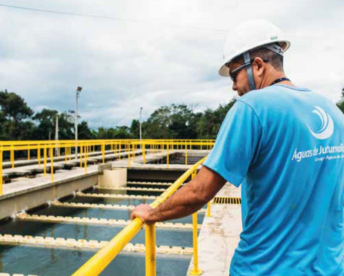 Homem com camiseta azul da Águas do Brasil e capacete de proteçao em planta de saneamento. Ele está com as mãos apoiadas em grade de amarela em torno da estação de tratamento.