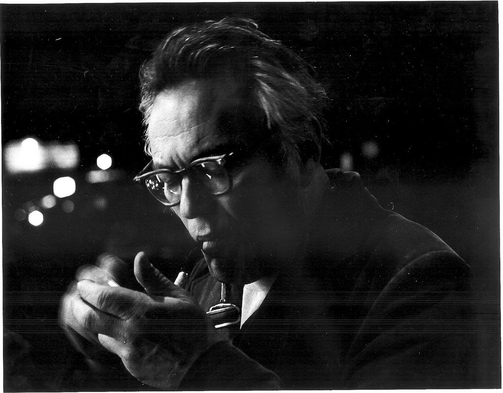 A middle-aged man wearing dark-framed glasses, with hair swept back from his broad forehead, cups his hands together as he lights a pipe; his right hand is blurred by motion. Behind him, out-of-focus streetlights arc back into the Manhattan night.