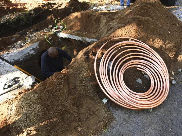 A worker is below street level replacing a lead water pipe. Dirt surrounds the top of the hole and new piping is coiled on the street. A worker is below street level replacing a lead water pipe. Dirt surrounds the top of the hole and new piping is coiled on the street.