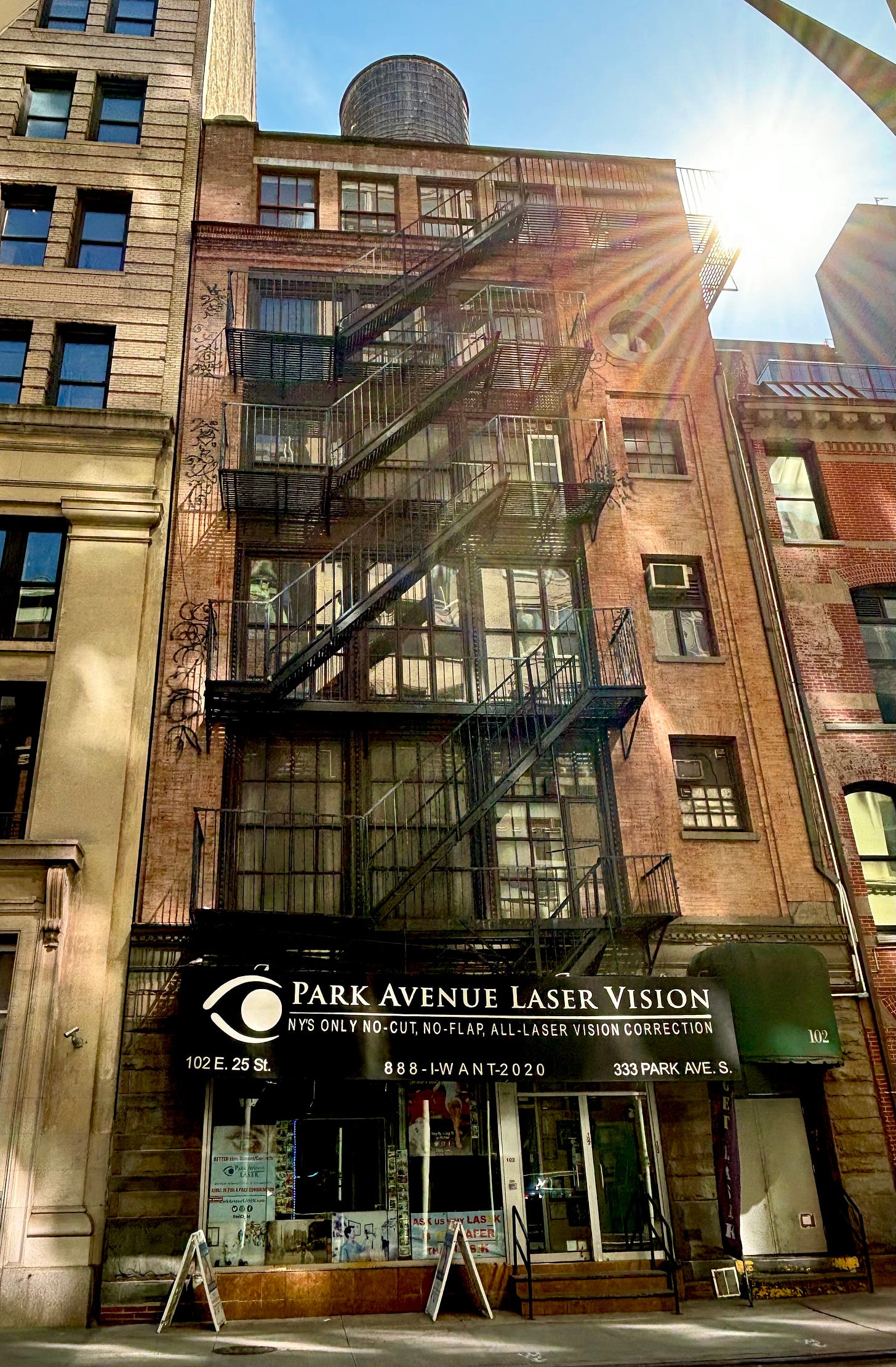 A five story brick building. There is graffiti running down the left side of the windows. On the second highest floor, there is a round window. The storefront has a black awning reading Park Avenue Laser Vision. NY'S Only No-Cut, No-Flap, All-Laser Vision Correction. The sun is blaring from the upper right corner and the rays are visible in front of the brick facade.
