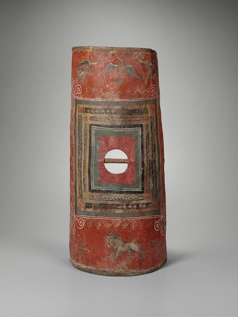 An amazing red barrel shield, a wonderful survivor. It’s decorated with stirring symbols of victory including eagles. You could form an impressive shield wall or ‘tortoise’ with this long oblong shield. The shield is bright brick red with squares of different colours in its centre — yellow, green, blue. It almost has a sort of no exit sign in its centre — ‘you shall not pass?’