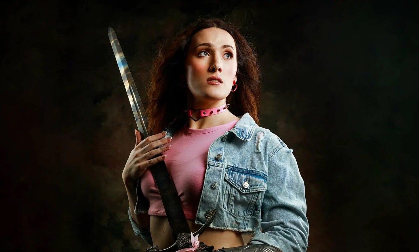 Photo of Abigail Thorn as a promotion for The Prince. She's a white woman wearing a pink crop top and cropped jean jacket holding a medieval sword. 