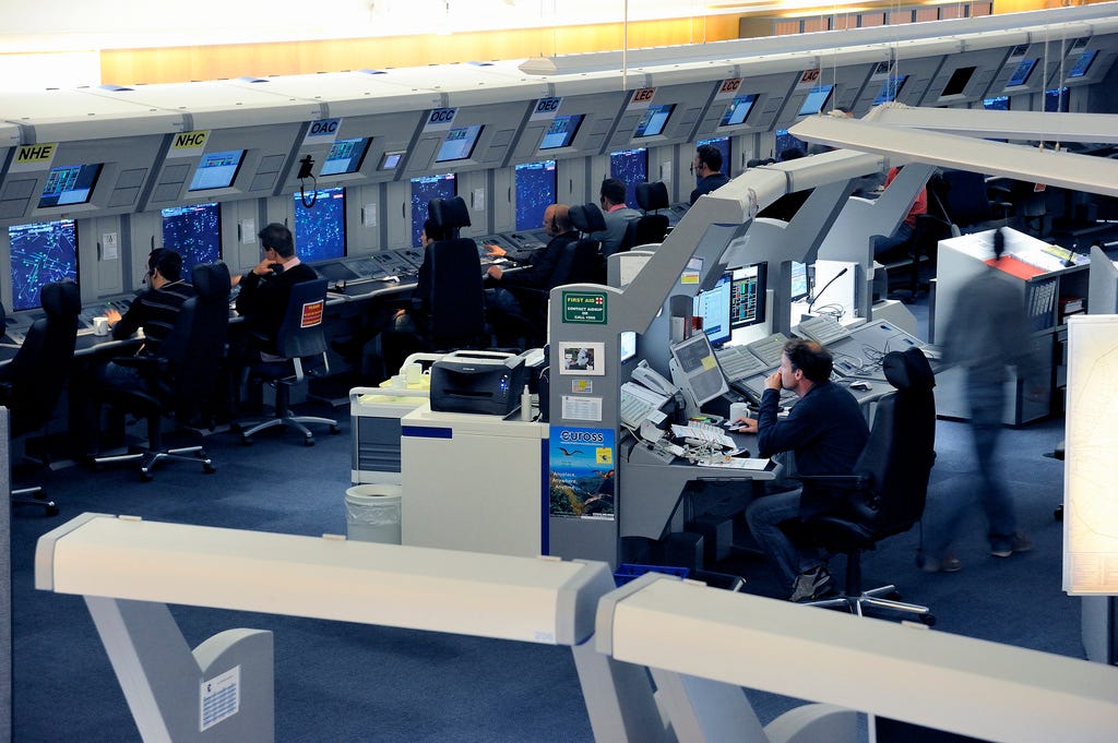 An air traffic control room showing two rows of computer screens staffed by attentive controllers. On each of their desks are two tiers of computer screens each showing complex graphics on blue screens. An air traffic control room showing two rows of computer screens staffed by attentive controllers. On each of their desks are two tiers of computer screens each showing complex graphics on blue screens.