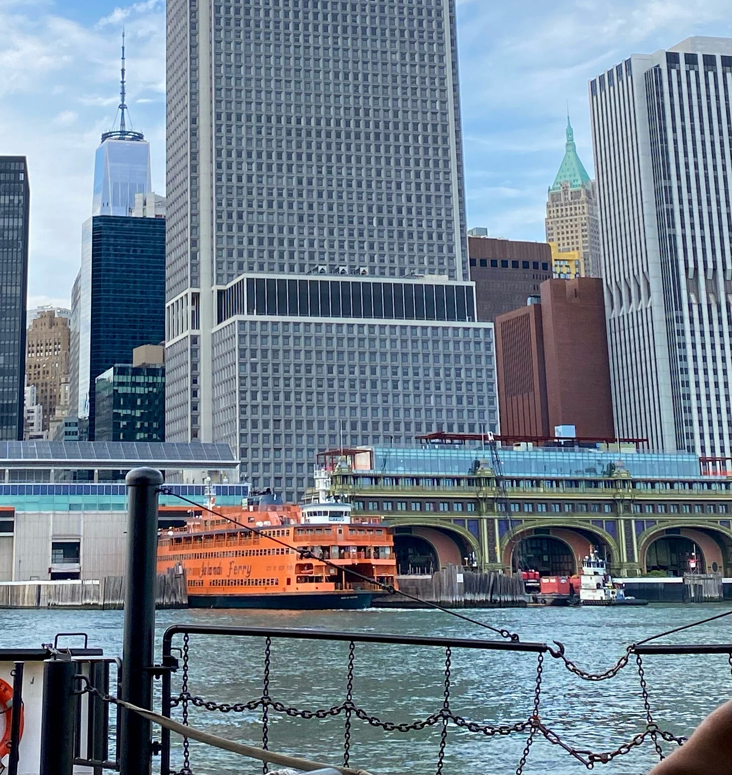 An orange Staten Island ferry pulls out of a terminal. Next to it is a more ornate, old fashioned terminal with three large arches. It is green with gold trim. Behind it are the skyscrapers of lower Manhattan with the WTC visible in the distance. The chain rail of a ferry and the East River are in the foreground.