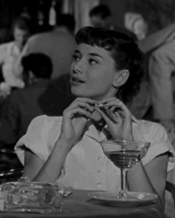 A GIF of 20th Century actress, Audrey Hepburn in an old movie looking coy at a dinner table and blowing a straw out of their mouth. 