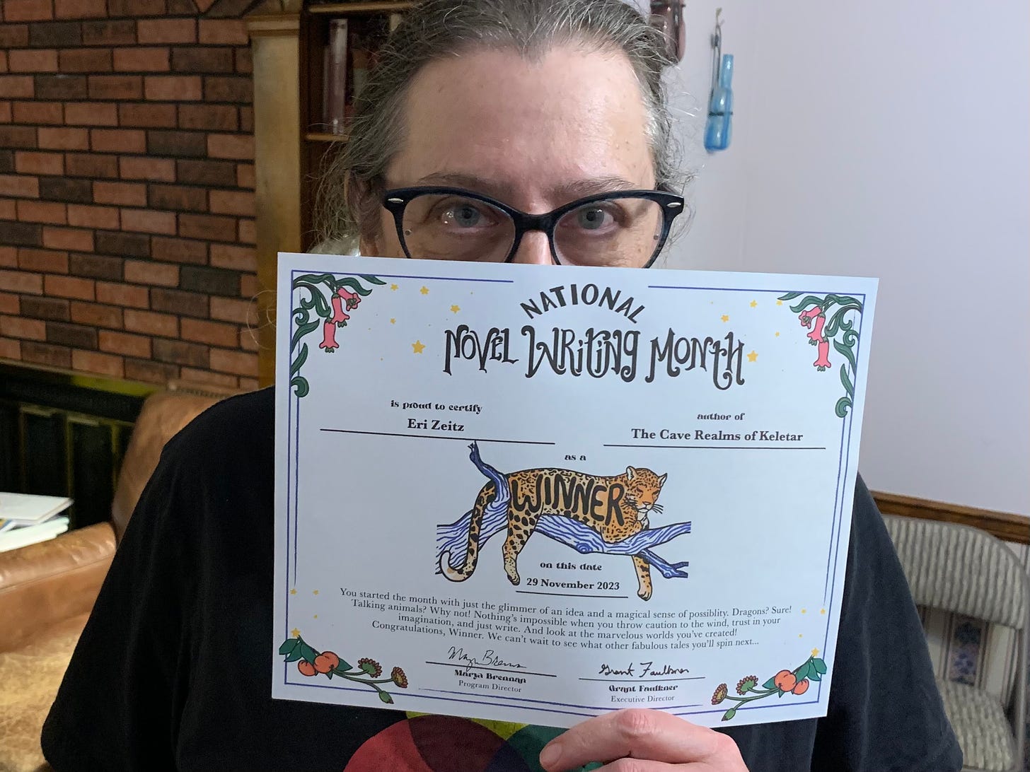 a woman with glasses holds a certificate in front of her:National Novel Writing Month. A leopard on a tree limb has the word WINNER on it's side.