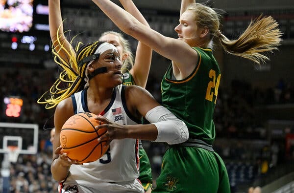 Aaliyah Edwards, at left in a white UConn jersey, holds the ball while Vermont’s Anna Olson, in a green jersey, raises her arms over Edwards.